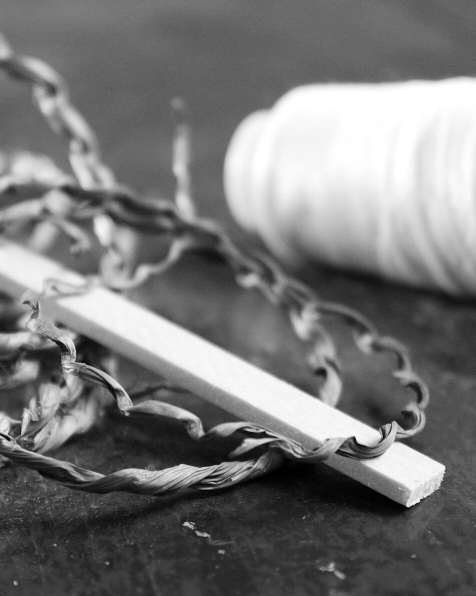 Black and white close-up photograph of natural crafting materials featuring a linden wood stick intertwined with twisted seaweed strands in the foreground, with a blurred white spool of polypropylene thread visible in the background.