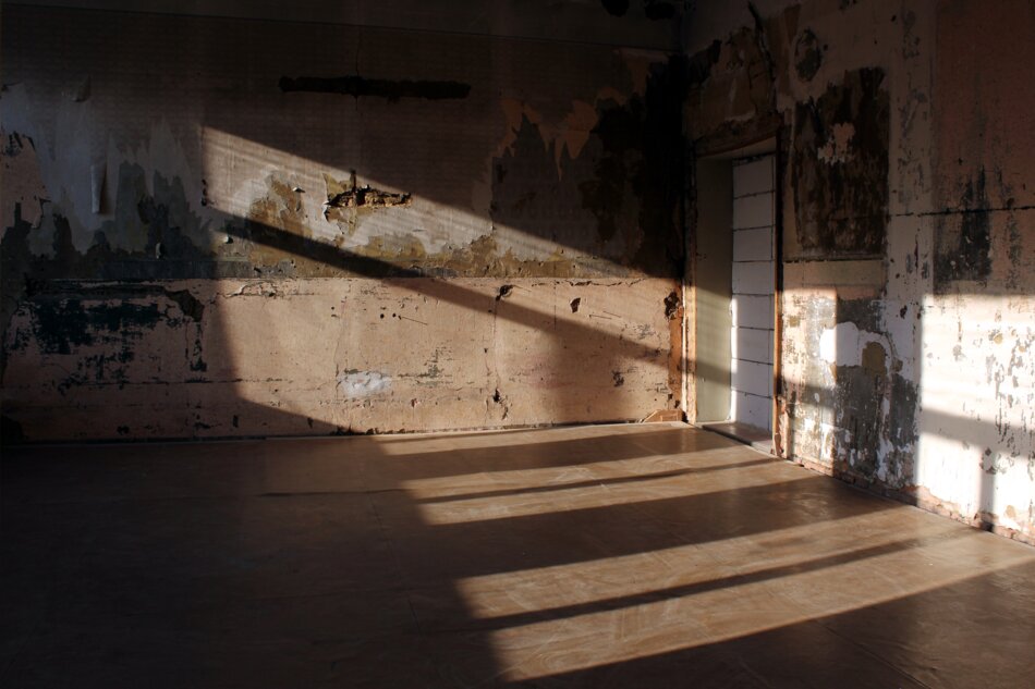 An abandoned room with peeling wallpaper and a broken door. Sunlight shines through the broken windows, casting long shadows on the floor.