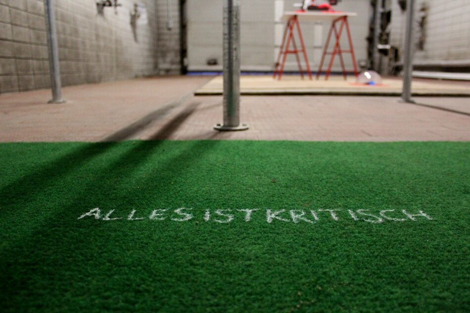 A photo of an industrial space with a concrete floor and tiled walls. A roll of green artificial turf lies on the floor with the German phrase “ALLES IST KRITISCH” written in white chalk across it. In the background, there is a red sawhorse, a metal pole, and a sink area with faucets.