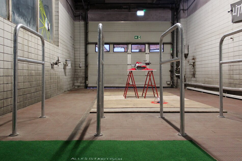An empty industrial space with a red table holding a typewriter in the center. The room features white tiled walls, metal railings, and a large metal door. The text “ALLES IST KRITISCH” is written on the floor.