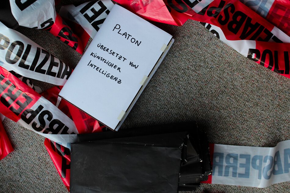 A crime scene photo featuring a white notebook labeled “PLATON ÜBERSETZT VON KÜNSTLICHER INTELLIGENZ” surrounded by red and white police tape.