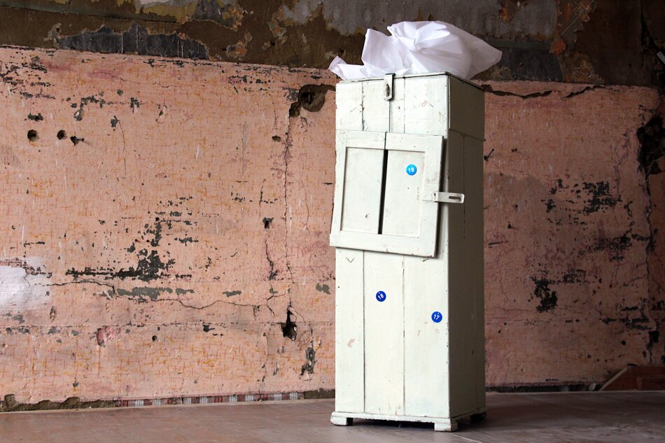 A white, weathered cabinet with a single door ajar stands in a room with peeling, pink-colored walls.