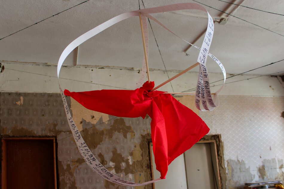 A hanging installation featuring long strips of paper with the repeated name “Heinz-Werner,” a red fabric element, and wooden structures.