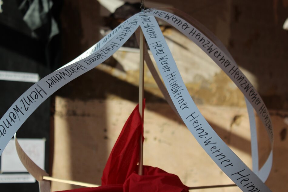 A hanging installation featuring long strips of paper with handwritten text, a red fabric element, and wooden structures.
