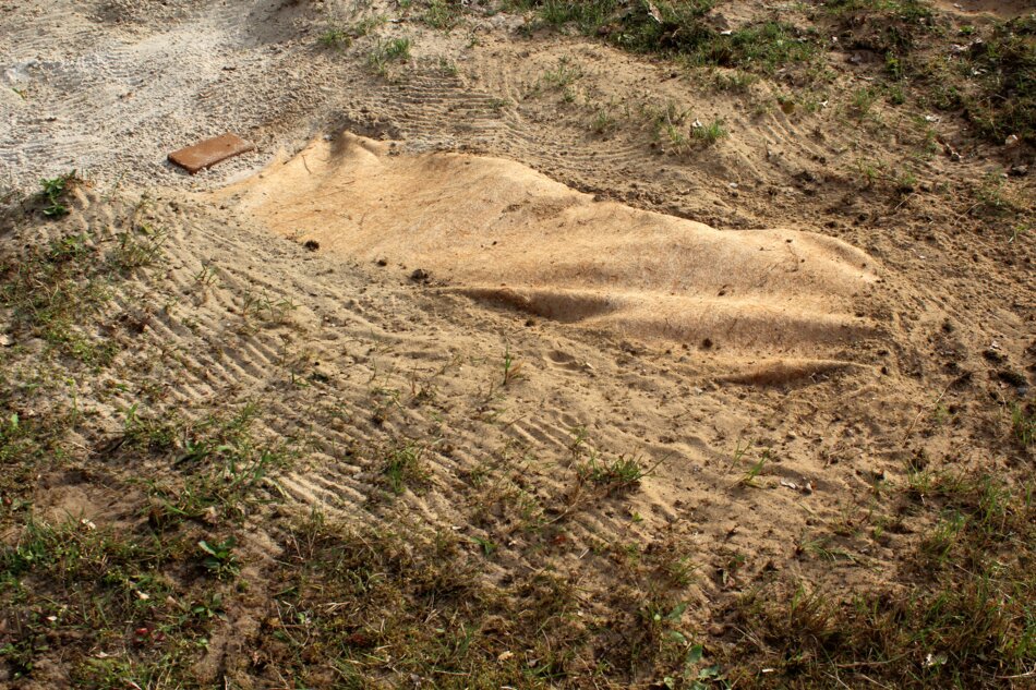 A pile of sand partially covered by a brown cloth, resting on a grassy area with tire tracks and a red brick in the background.