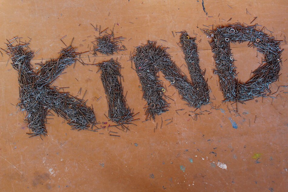 The word “KIND” is spelled out in nails on a brown surface. The background is cluttered with paint splatters.