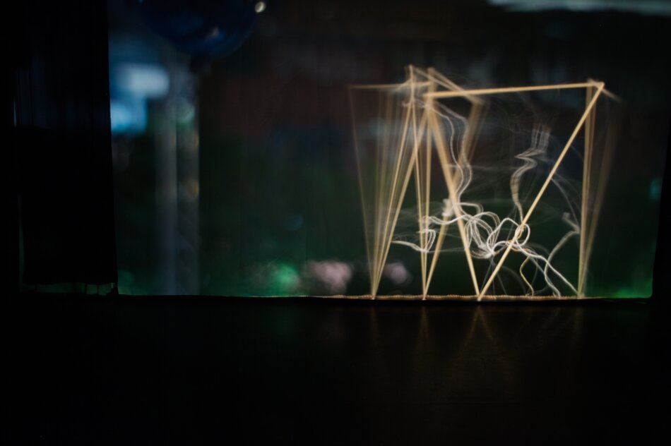 A projection in a human-scale camera obscura of an object made of four linden strip triangles, seaweed threads, and polypropylene fibers. The object appears partially blurred due to movement, with a bokeh effect in the background.