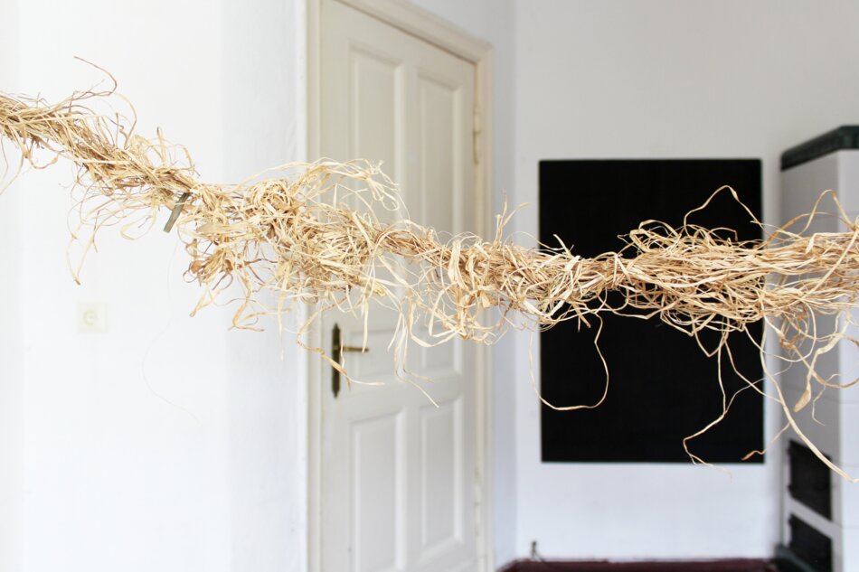 A close-up detail of a raffia artwork hanging in a room with white walls,
 a wooden door, a black chalkboard, and a white stove. The raffia is arranged in a loose, flowing manner, creating a sense of movement and texture.