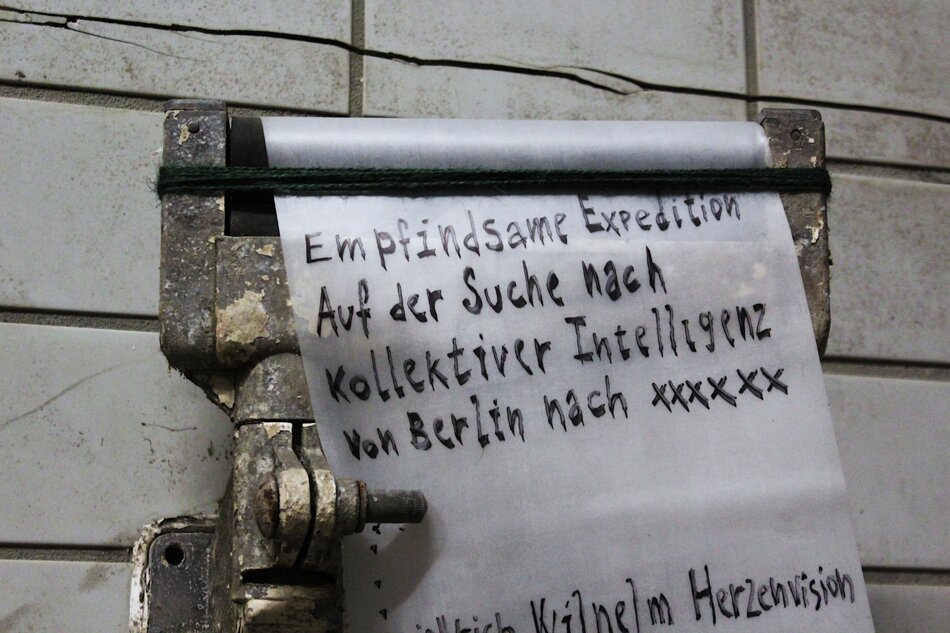 Black and white photo of a weathered silicon sheet mounted on a rusty metal device, hanging against a tiled wall. The sheet contains handwritten text in German: “Empfindsame Expedition Auf der Suche nach Kollektiver Intelligenz von Berlin nach xxxxxx” and a signature.