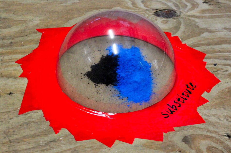 A transparent dome sits on a red, star-shaped base with the word “Substitute” written on it. Inside the dome, black and blue powder is divided into two sections. The entire setup is placed on a wooden surface.