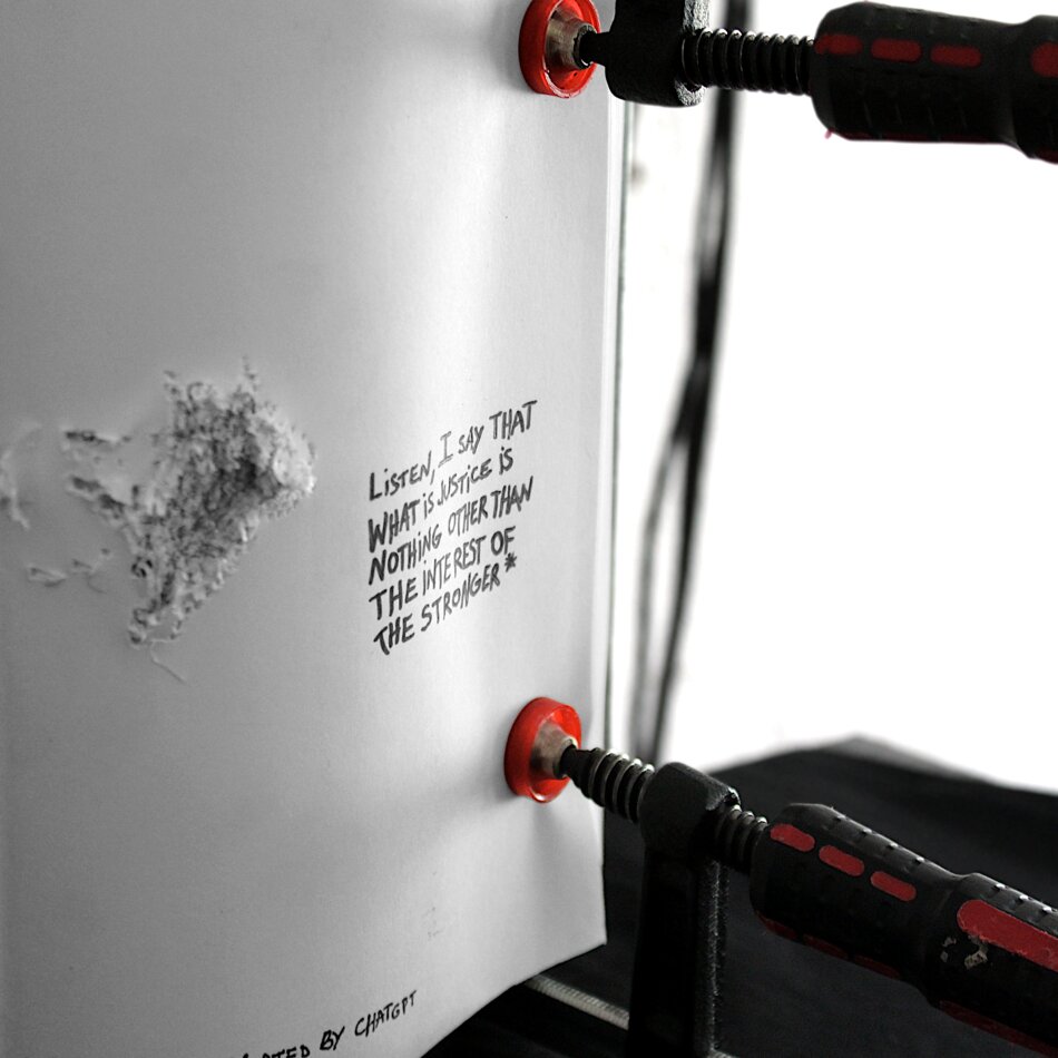 A white sheet of paper with a bullet hole in the center, surrounded by a dark stain. Handwritten text in black ink reads “LISTEN, I SAY THAT WHAT IS JUSTICE IS NOTHING OTHER THAN THE INTEREST OF THE STRONGER” with a signature below. The paper is clamped between two red and black mechanical clamps.