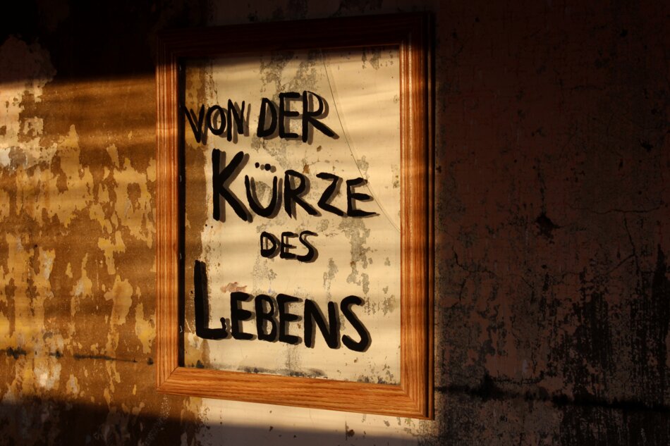 A framed mirror with the text “VON DER K&Uuml;RZE DES LEBENS” written in black marker on it. The mirror is hanging on a weathered wall with peeling paint.